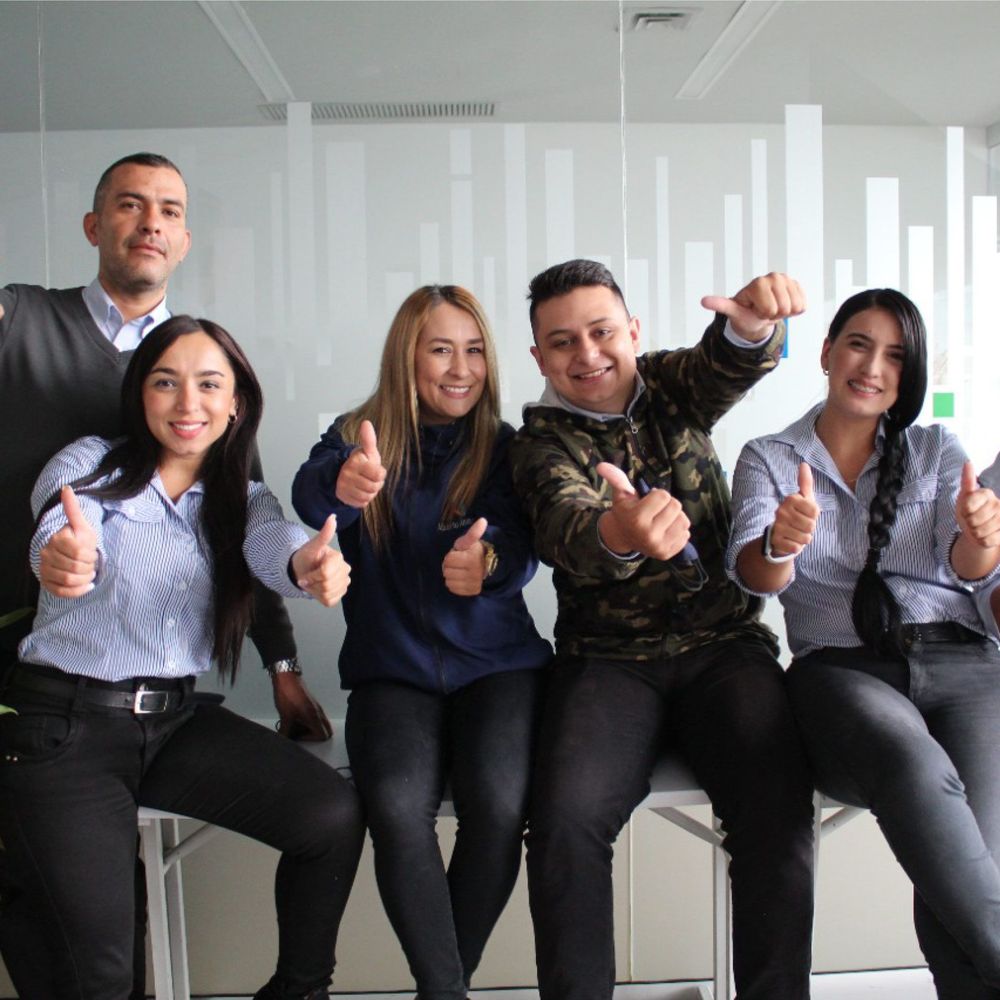 A group of six smiling young adults gives a thumbs up to the camera.