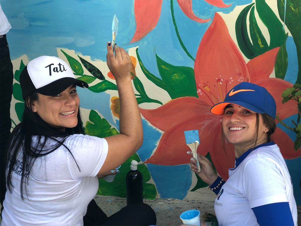 Two smiling women paint a colorful mural with large floral designs.